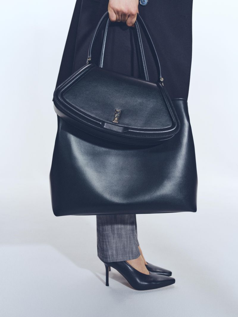 Elegant black leather tote bag held by a person wearing grey slacks and black high heels against a plain white backdrop.