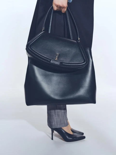 Elegant black leather tote bag held by a person wearing grey slacks and black high heels against a plain white backdrop.