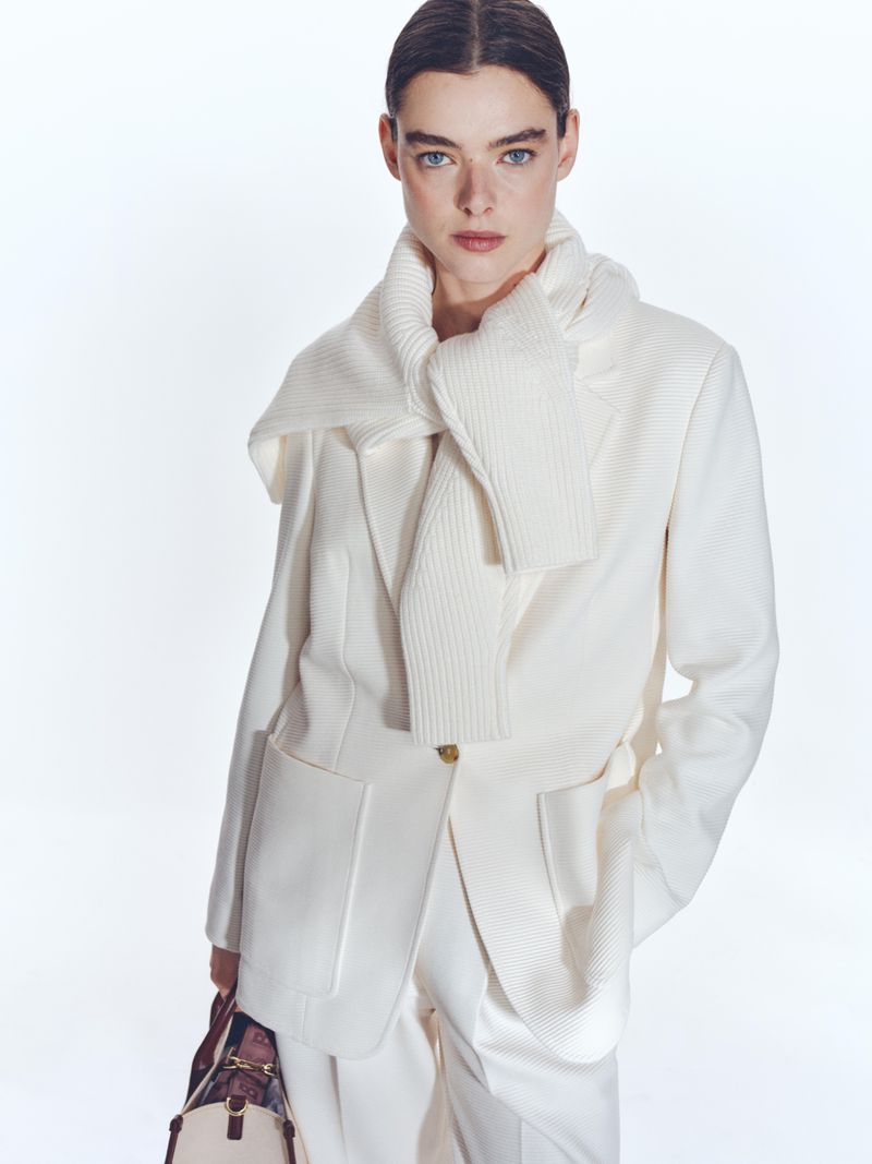 Fashion editorial shot of a woman wearing an off-white blazer and scarf, and carrying a satchel-style purse, against a white background.