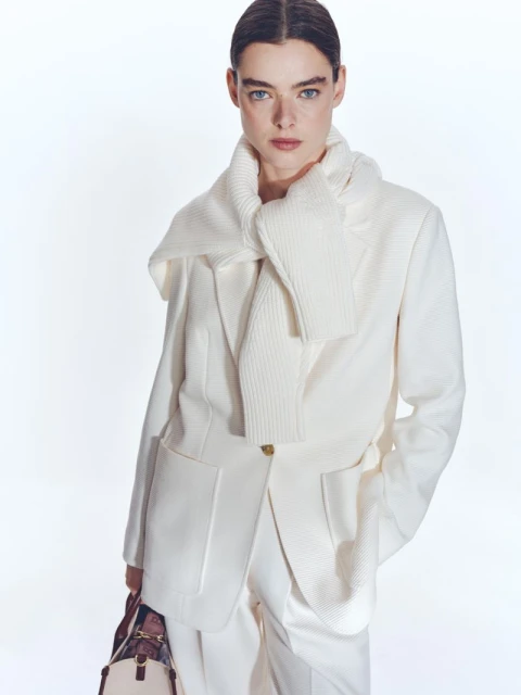 Fashion editorial shot of a woman wearing an off-white blazer and scarf, and carrying a satchel-style purse, against a white background.