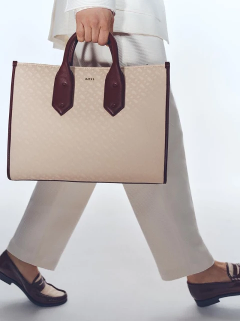 Walking woman carrying a cream-colored tote handbag with dark brown trim and handles, wearing a cream suit and matching loafers.
