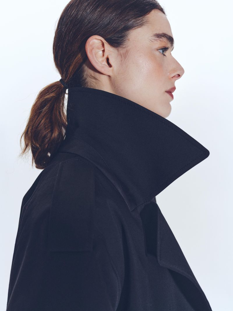 Profile shot of a woman with brown hair pulled back, wearing a black coat with a wide collar against a clean white background.