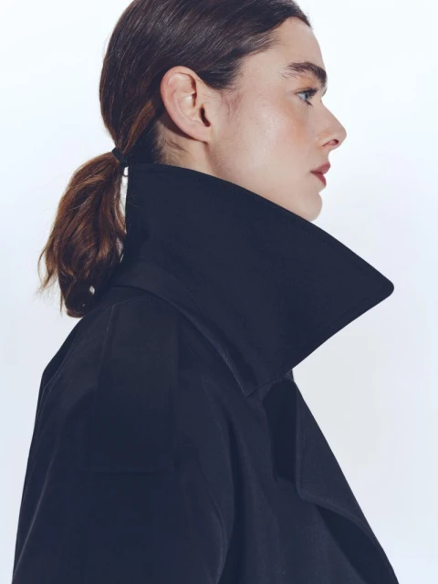 Profile shot of a woman with brown hair pulled back, wearing a black coat with a wide collar against a clean white background.