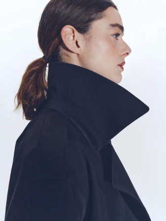 Profile shot of a woman with brown hair pulled back, wearing a black coat with a wide collar against a clean white background.
