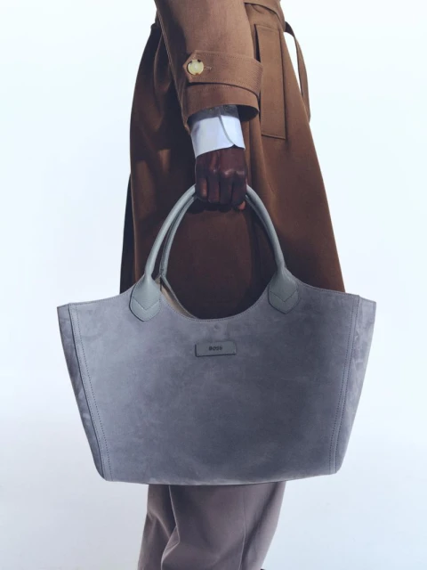 Dark-skinned hand holding a large grey suede tote bag, tan overcoat, and white studio background in a fashion photography setting.