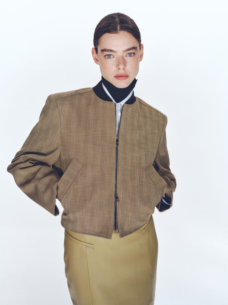 Woman in brown jacket and tan skirt poses in front of white background in studio setting. Fashion editorial shot. Beauty shot of young model.