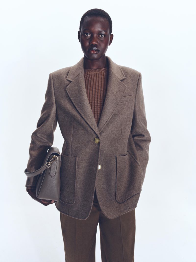 Woman in a brown blazer, dark sweater, and brown pants, carrying a gray handbag against a bright white background in a studio shot.