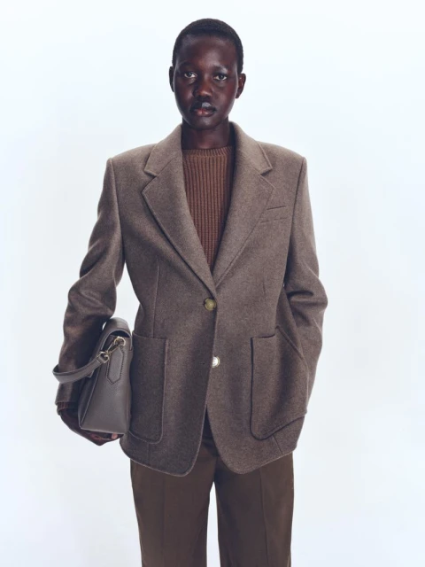 Woman in a brown blazer, dark sweater, and brown pants, carrying a gray handbag against a bright white background in a studio shot.