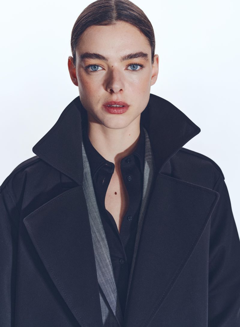 Portrait of a woman with blue eyes and dark hair wearing a dark trench coat against a white background in a fashion forward style.