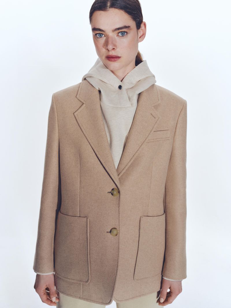 Model wearing a beige tailored blazer over a hooded sweatshirt against a white background