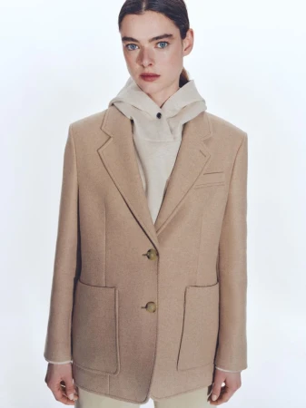 Model wearing a beige tailored blazer over a hooded sweatshirt against a white background