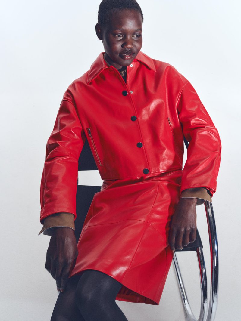 Model wearing a red leather jacket and matching skirt seated on a chrome chair against a white background for BOSS