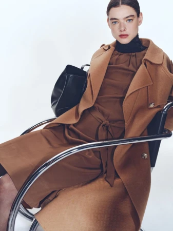 Model seated in a chrome armchair wearing a camel wool coat over a brown dress and black leather handbag
