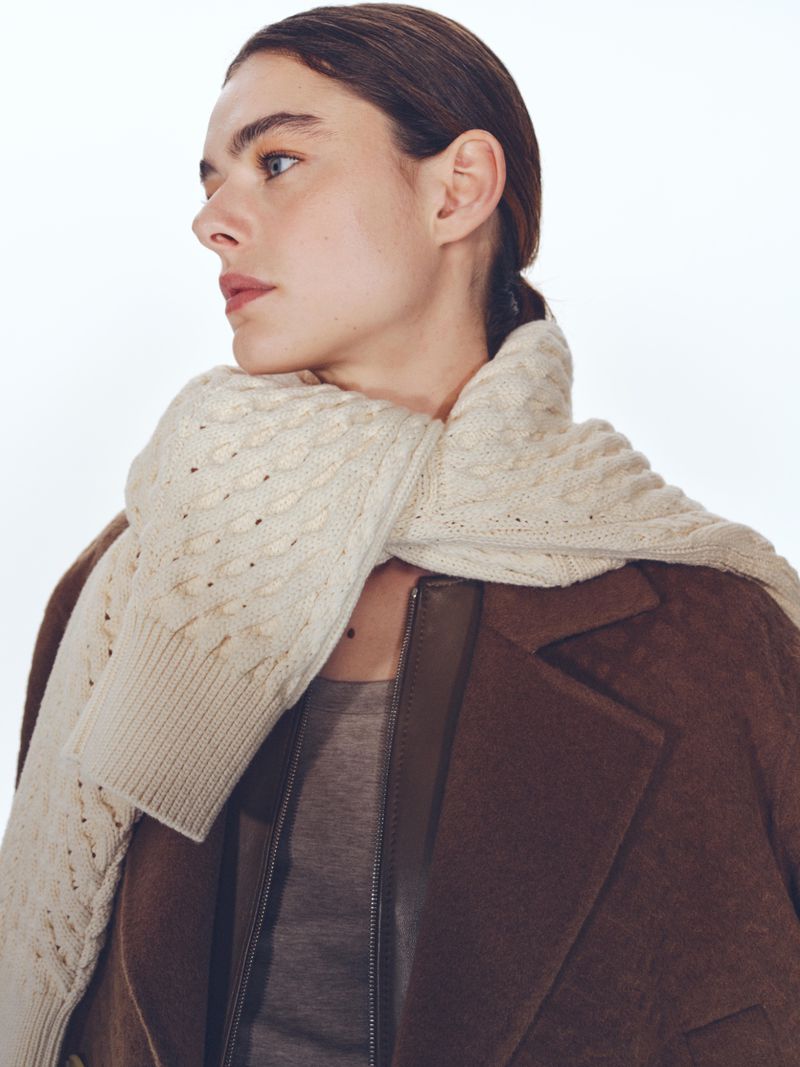 Portrait of a woman wearing a brown coat and cream scarf in a bright, neutral studio setting. Focus on fashion and soft, diffused lighting.