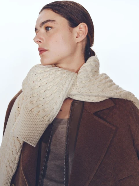 Portrait of a woman wearing a brown coat and cream scarf in a bright, neutral studio setting. Focus on fashion and soft, diffused lighting.