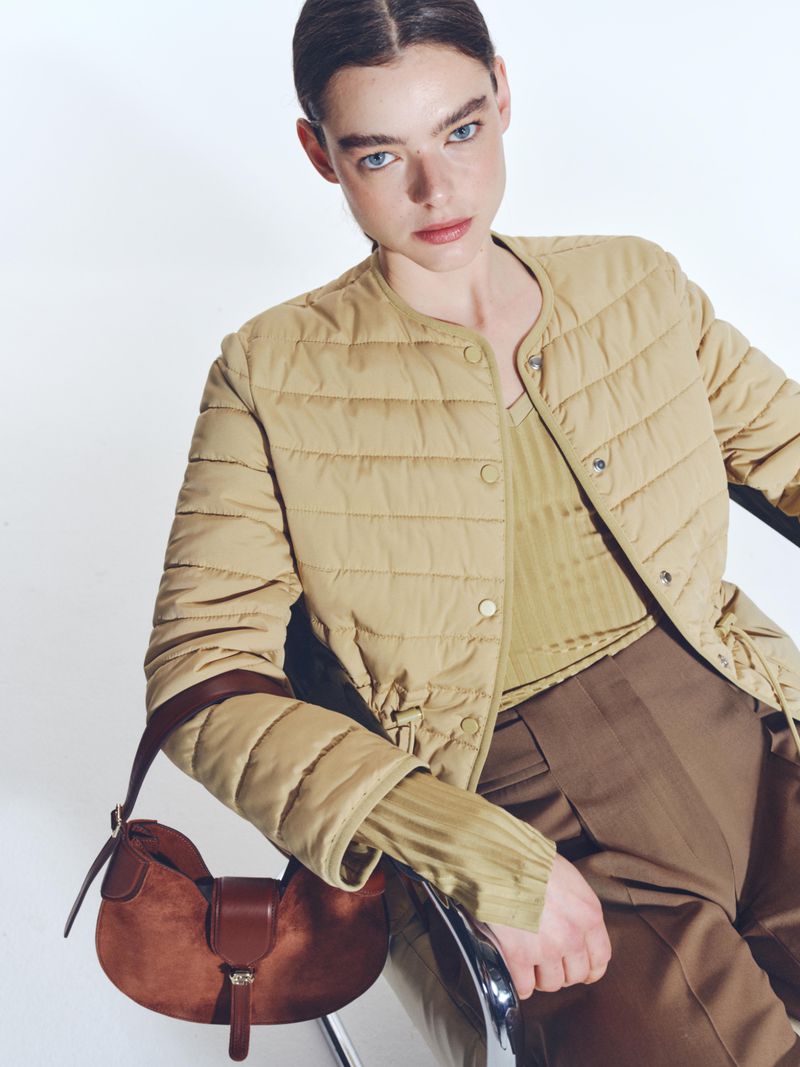 Fashion shot of a woman wearing a beige quilted jacket, brown pants, and a brown shoulder bag, set against a plain white backdrop.