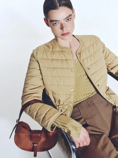 Fashion shot of a woman wearing a beige quilted jacket, brown pants, and a brown shoulder bag, set against a plain white backdrop.