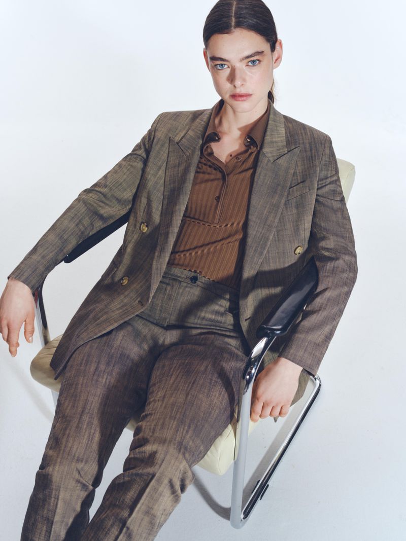 An adult female model poses in a brown suit, seated in a metal chair against a white backdrop, presented in a medium studio shot.