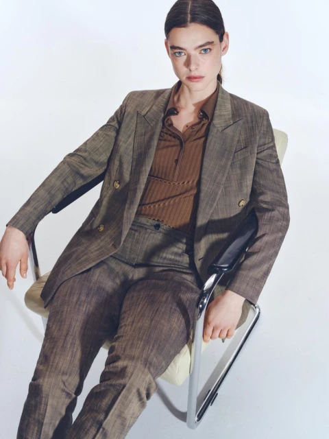 An adult female model poses in a brown suit, seated in a metal chair against a white backdrop, presented in a medium studio shot.