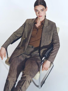 Model seated in an armchair wearing a brown tweed suit and brown shirt against a white studio background