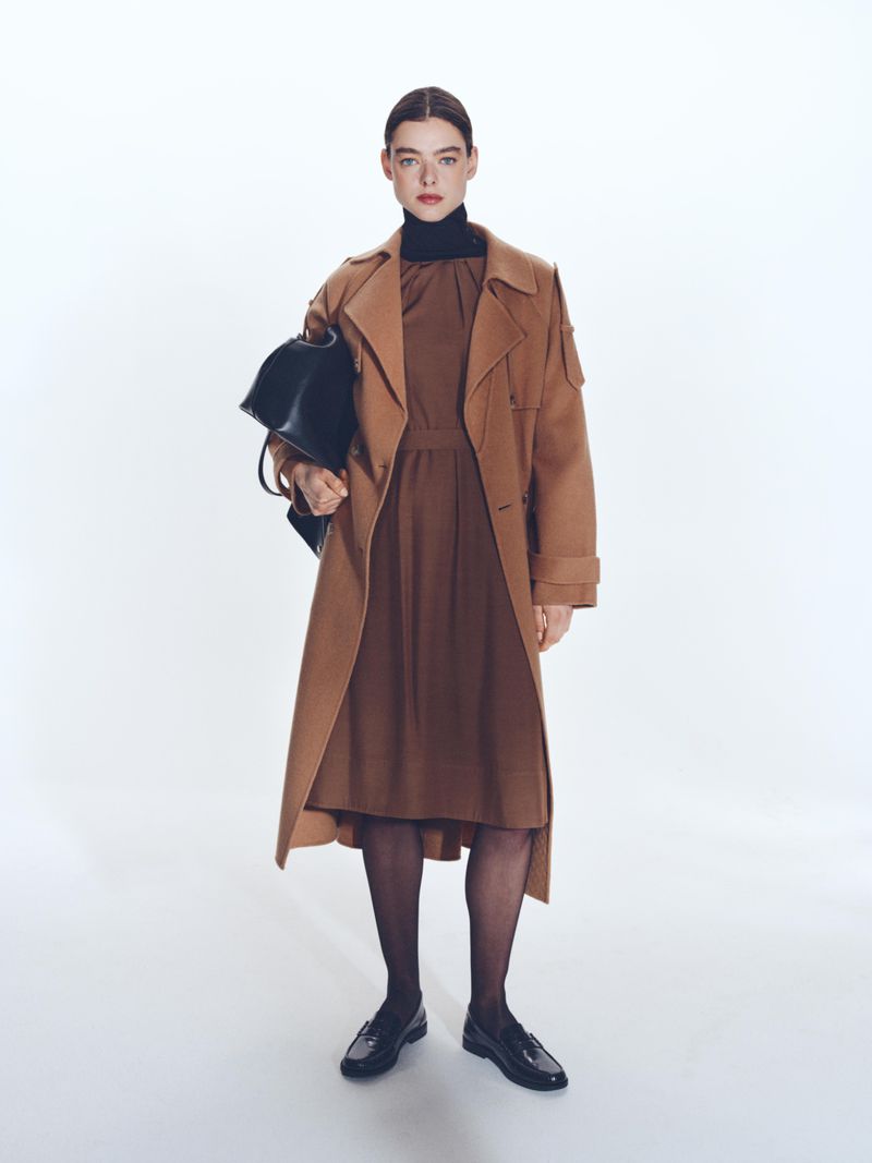 Model wearing a camel trench coat and brown dress holding a black handbag against a white background for BOSS Spring 2025.