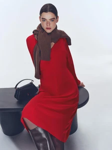 Model in a red dress with brown scarf and boots sitting on a black bench against a seamless white background.