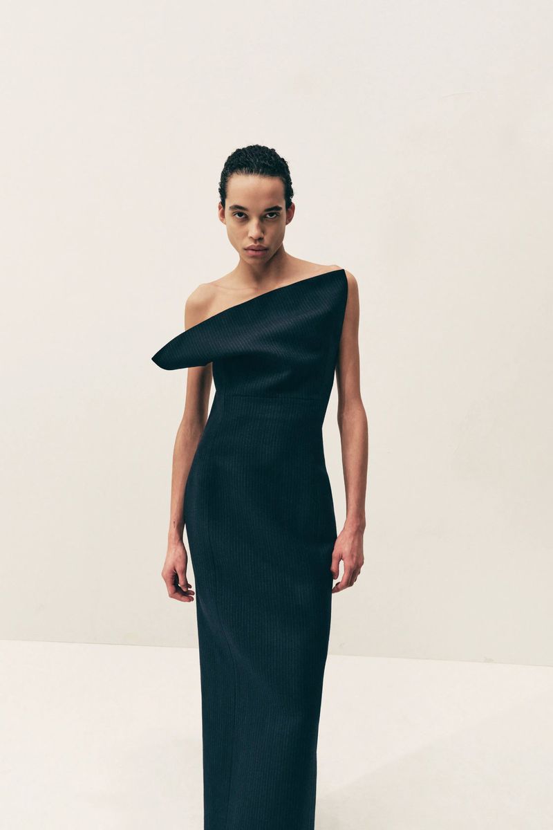 Elegant woman poses in a black, one-shoulder maxi dress with a sleek, straight hairstyle against a soft white backdrop in a fashion-forward shot.