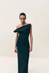 Model in a structured black off-the-shoulder dress standing against a plain pale studio background.