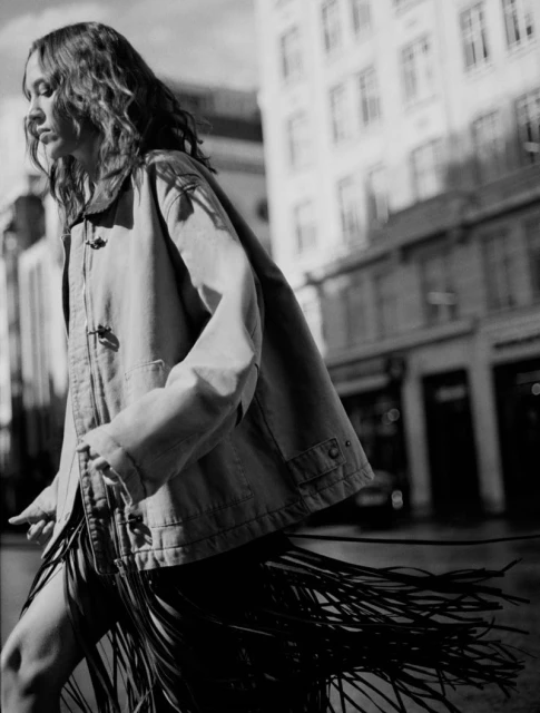 A woman walks in a city, wearing a jacket and a fringe skirt, captured in a black and white medium shot with blurred background.