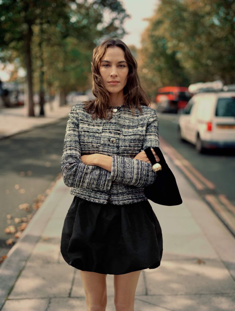 Adult female in a tweed jacket and black skirt stands on a city street with trees and vehicles in background, holding a small black bag.