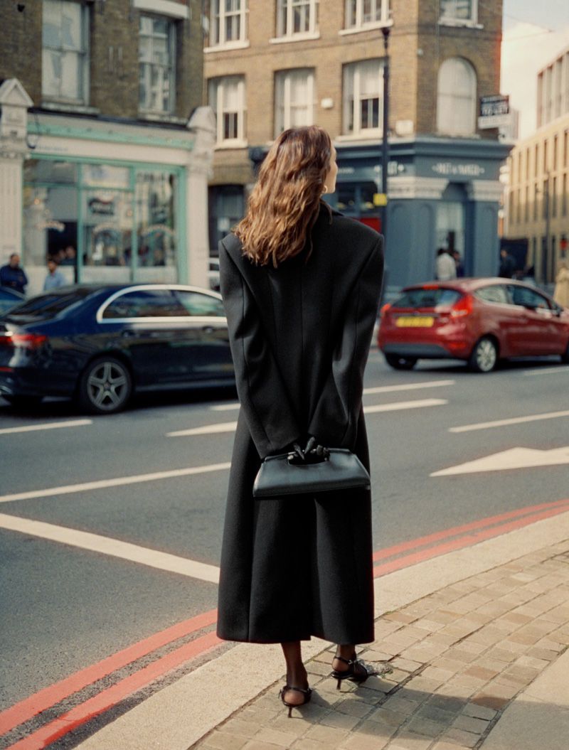 Woman in a long black coat holding a black handbag standing on a city street