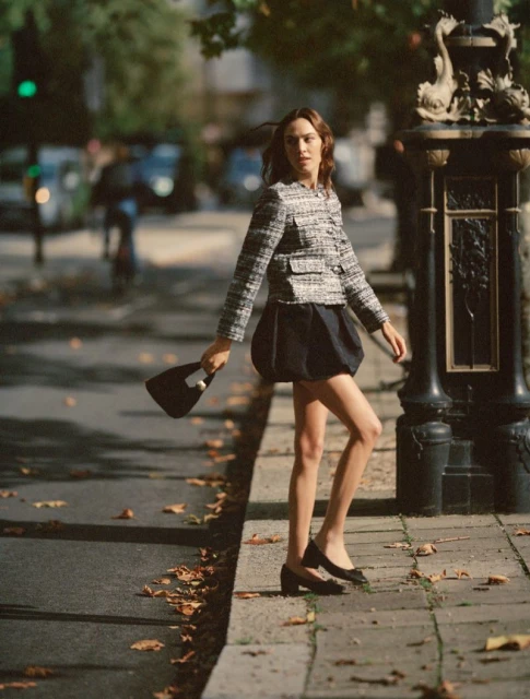 Elegant adult female in a tweed jacket, navy skirt, and black heels walking on a city sidewalk with fallen leaves, holding a small handbag.
