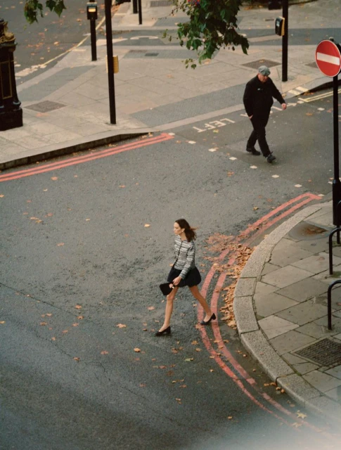 Woman in a striped shirt and skirt crossing a street in an urban setting as a man crosses in the background, captured in daylight.