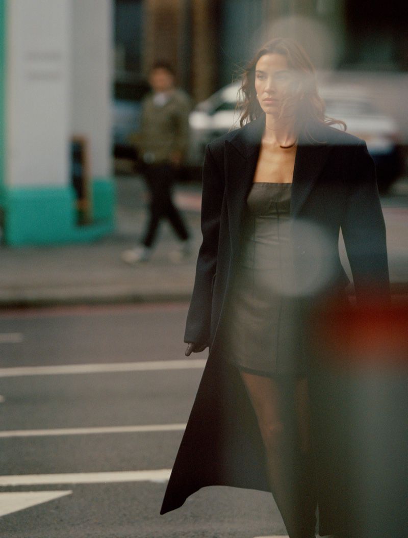 A woman in a black coat walking on a city street with blurred pedestrians and cars in the background, creating a shallow depth of field.
