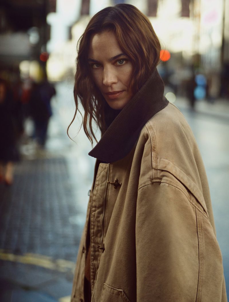 An adult woman with long brown hair poses in an urban street wearing a brown workwear jacket and looking directly at the camera.
