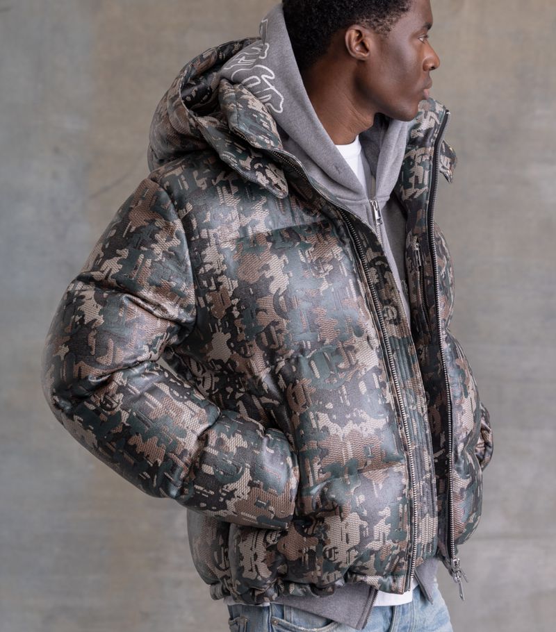 Profile shot of an adult male wearing a camouflage-patterned puffer jacket, gray hoodie, and jeans against a neutral backdrop.