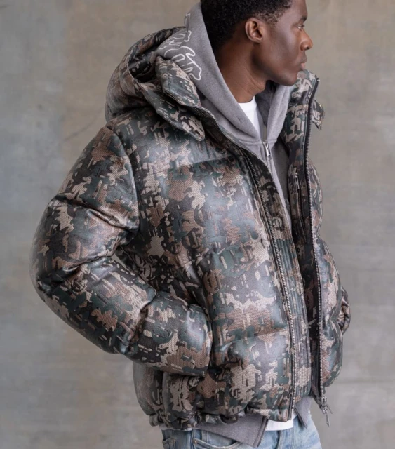 Profile shot of an adult male wearing a camouflage-patterned puffer jacket, gray hoodie, and jeans against a neutral backdrop.