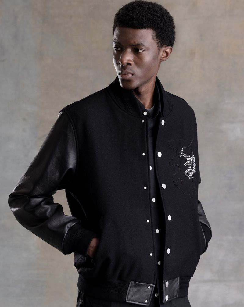 A stylish young Black man models a black varsity jacket with leather sleeves against a mottled grey backdrop in a medium shot.