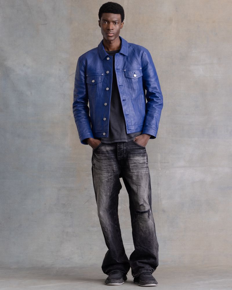 Portrait of a young Black man wearing a blue leather jacket, grey t-shirt and black denim pants, posing in a studio with a grey backdrop.