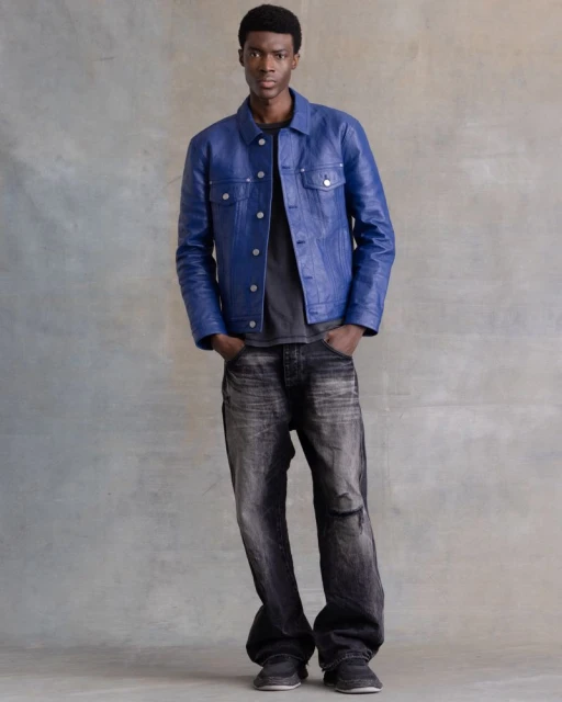 Portrait of a young Black man wearing a blue leather jacket, grey t-shirt and black denim pants, posing in a studio with a grey backdrop.