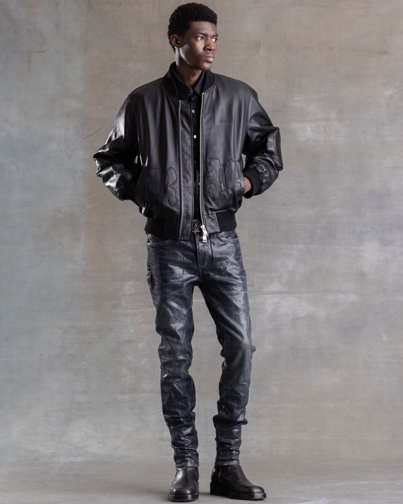 A Black man stands in a studio setting wearing a black leather bomber jacket, dark button-down shirt, glossy pants, and black boots.