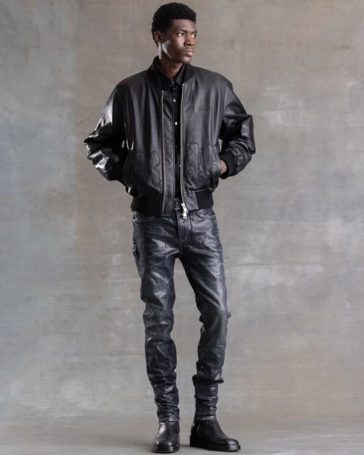 A Black man stands in a studio setting wearing a black leather bomber jacket, dark button-down shirt, glossy pants, and black boots.