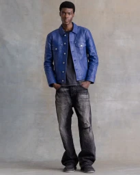Full-length portrait of a Black man wearing a royal blue leather jacket and dark distressed jeans standing against a grey textured studio backdrop.