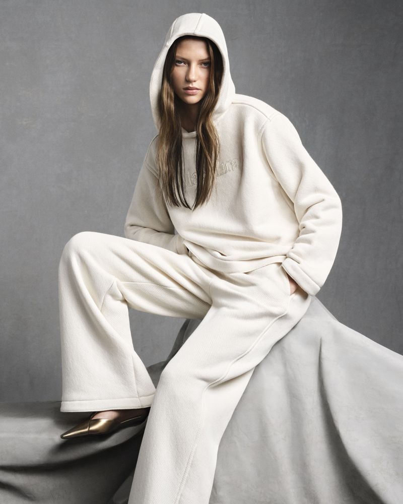 Fashion portrait of a young woman in a cream-colored hooded sweatshirt and wide-leg pants, seated on a grey fabric backdrop. Studio lighting.