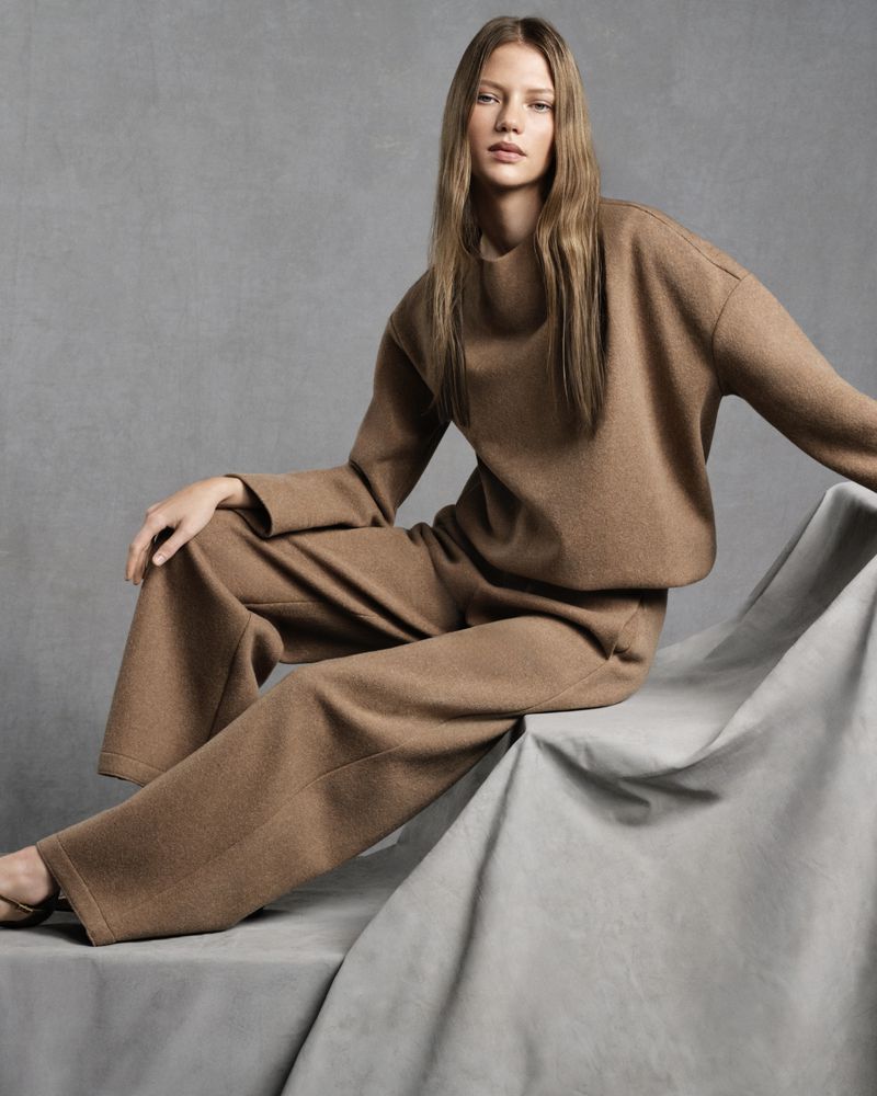 A woman is sitting gracefully on a draped surface, wearing a tan, long-sleeved sweater and wide-legged pants, indoor studio fashion shot.
