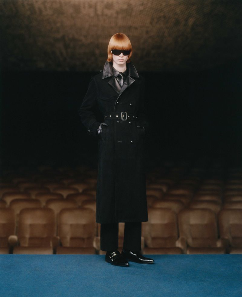 A redhead fashion model poses in a black leather-trimmed trench coat with sunglasses, standing against a dark theater backdrop.