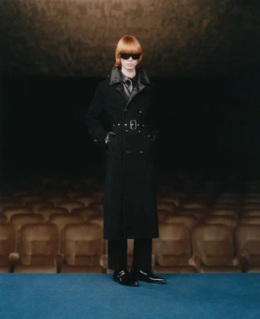 A redhead fashion model poses in a black leather-trimmed trench coat with sunglasses, standing against a dark theater backdrop.
