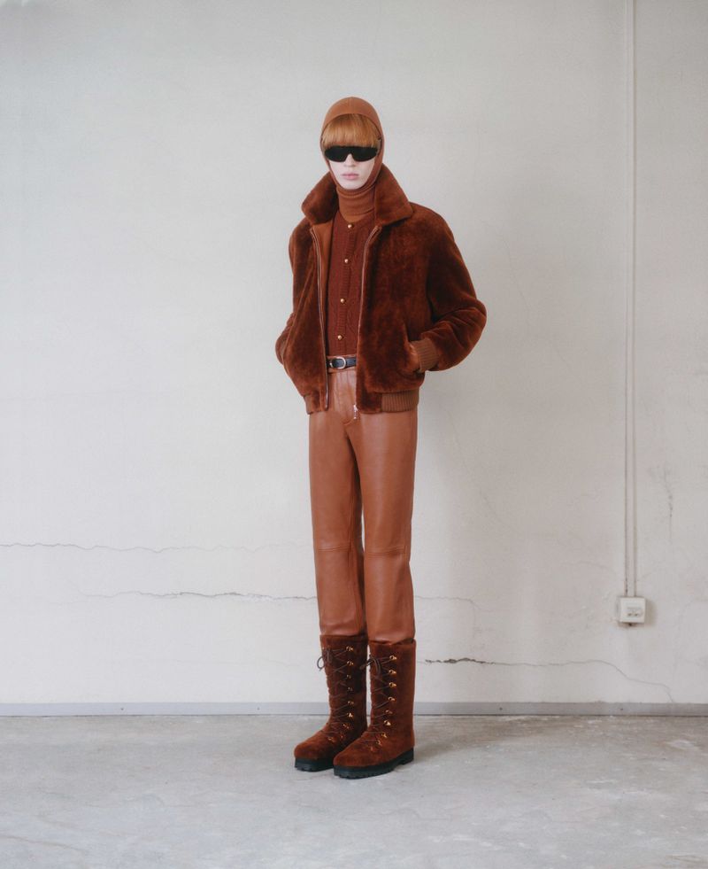 Male fashion model wears a brown fur coat, brown turtle neck, matching pants, beanie, sunglasses, and lace-up boots in a studio setting.