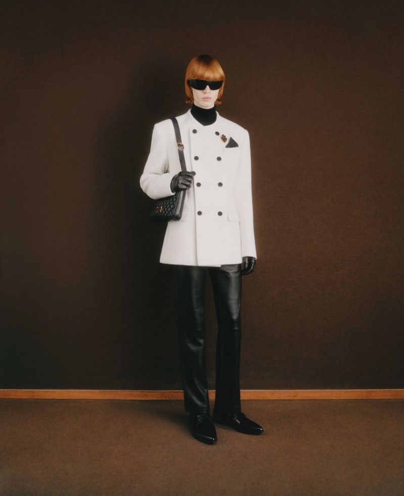 Full shot of a slender adult male model posing in a white double-breasted suit, black leather pants, and sunglasses against a brown backdrop.