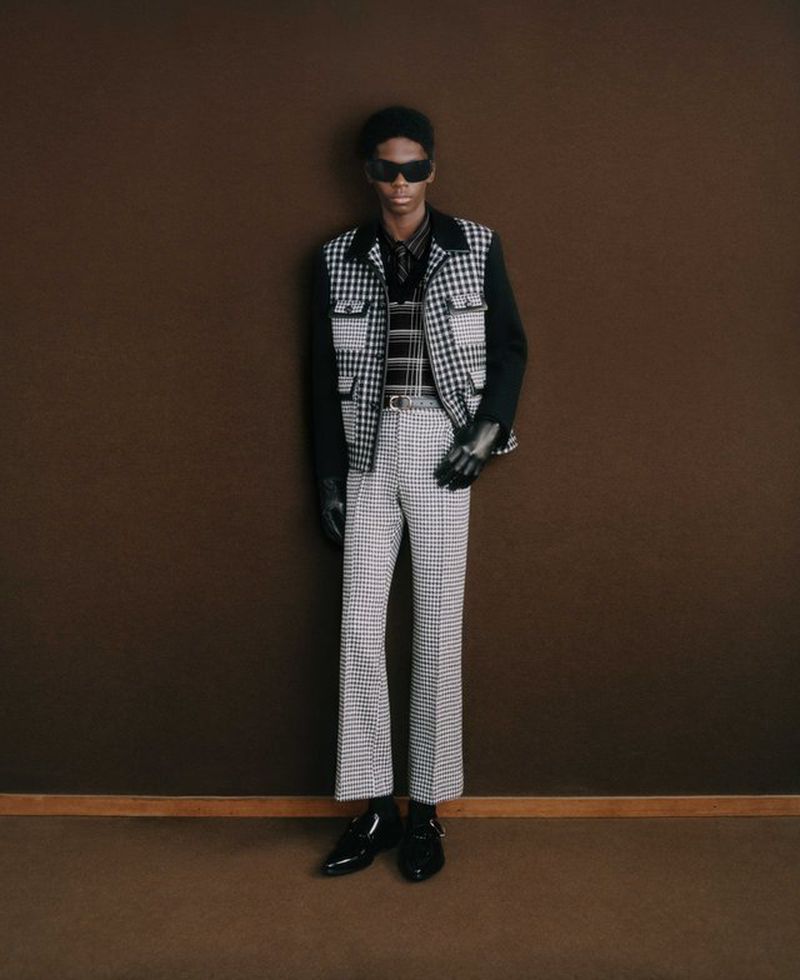 Fashionable African-American male model poses in black and white houndstooth vest and pants against a brown wall in a studio setting.